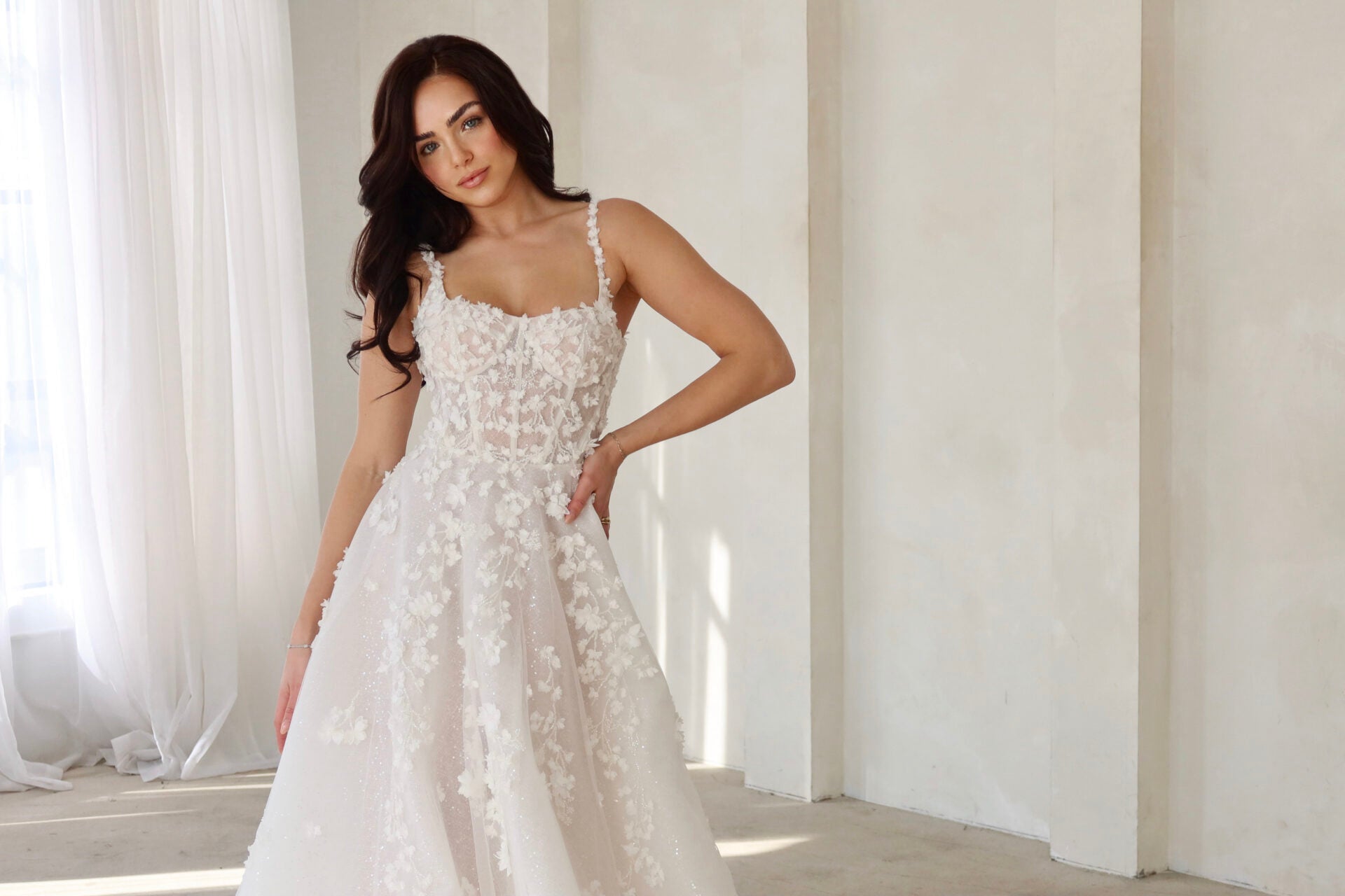 Woman wearing a white wedding dress in a softly lit room with white curtains.