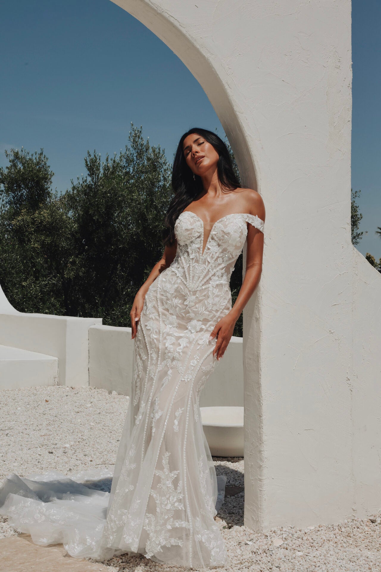Woman in a white wedding dress standing under a white archway with greenery in the background.