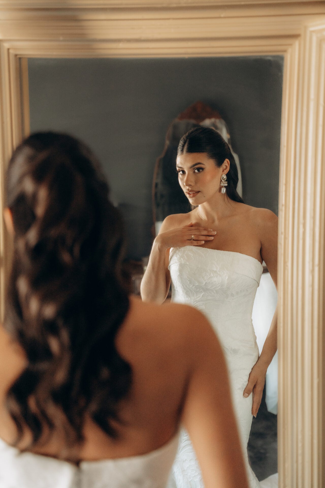 Woman in a white strapless dress looking at herself in a mirror.